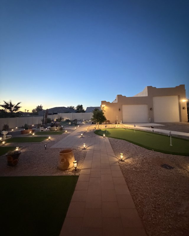 The same backyard at dusk after landscaping — lit paver walkways, turf panels, and a putting green