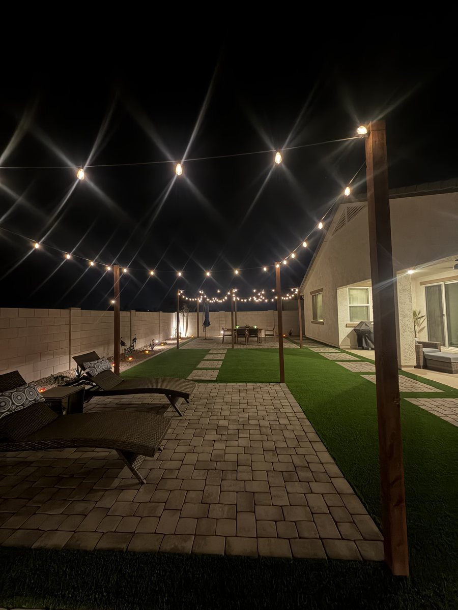 Bistro string lights strung on wood posts over a paver patio and artificial turf at night